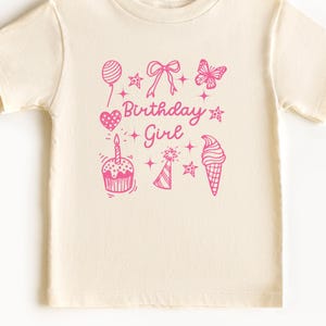 May include: A cream colored t-shirt with a pink graphic design that says "Birthday Girl" and includes a birthday cake, a party hat, a balloon, a heart, a butterfly, and an ice cream cone.