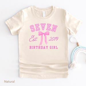 Peut inclure: Un t-shirt de couleur naturelle avec le texte rose "SEVEN", au-dessus d'un n&oelig;ud rose et du texte "Est. 2019 BIRTHDAY GIRL". Le t-shirt a des manches courtes et un col rond.