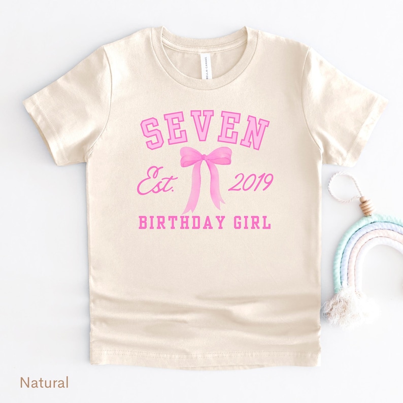 Peut inclure: Un t-shirt de couleur naturelle avec le texte rose "SEVEN", au-dessus d'un n&oelig;ud rose et du texte "Est. 2019 BIRTHDAY GIRL". Le t-shirt a des manches courtes et un col rond.