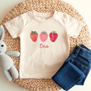 May include: A cream-colored toddler t-shirt featuring three strawberries in red and pink hues. The word "One" is written in a cursive font below the strawberries. Dark blue denim jeans are folded next to the shirt.