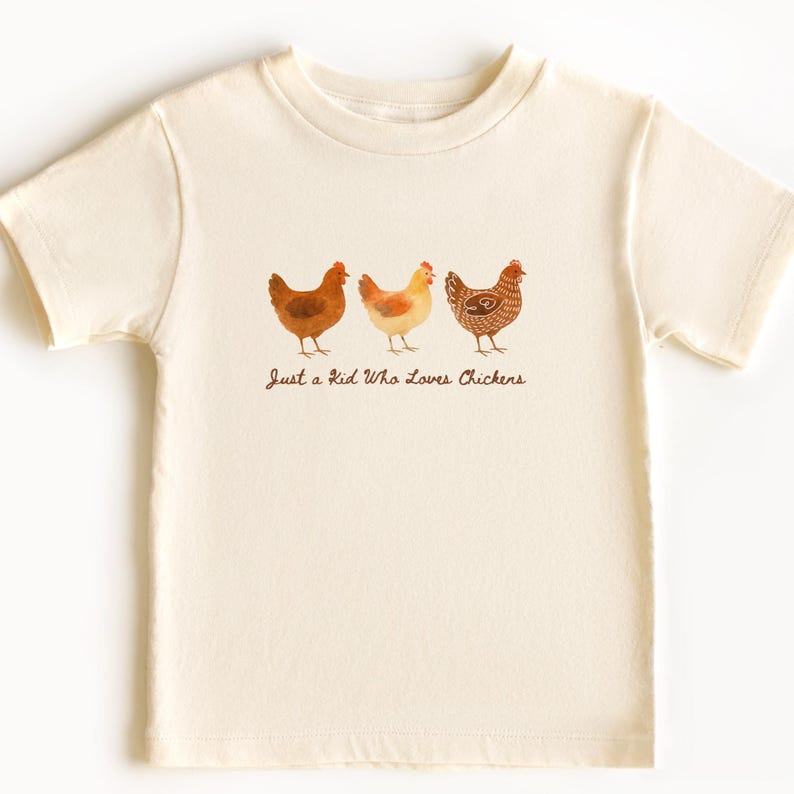 May include: Cream-colored t-shirt featuring three watercolor chickens in shades of brown and yellow. The text "Just a Kid Who Loves Chickens" is printed below the chickens. The shirt has short sleeves.