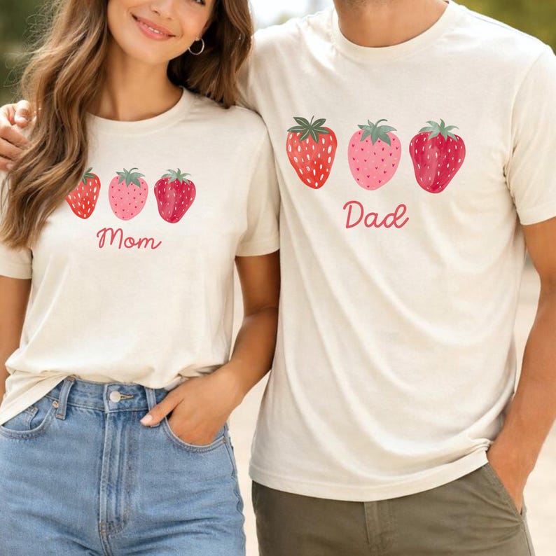 May include: Matching beige t-shirts with strawberry graphics. One shirt says "Mom" and the other says "Dad" in red script. The strawberries are red and pink with green leaves. The shirts are short-sleeved.