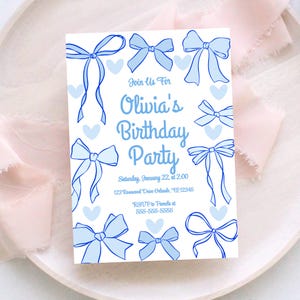 Puede incluir: Una tarjeta de invitación blanca con un diseño de lazo y corazón azul claro. El texto dice "Join Us For Olivia's Birthday Party" con fecha, hora, dirección e información de RSVP. La tarjeta está sobre un plato blanco con tela rosa.