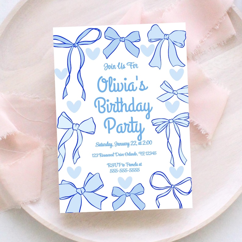May include: A white invitation card with a light blue bow and heart design. The text reads "Join Us For Olivia's Birthday Party" with date, time, address, and RSVP information. The card is on a white plate with pink fabric.