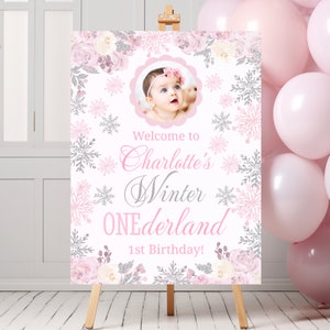 May include: A pink and silver welcome sign with a snowflake design. The sign says "Welcome to Charlotte's Winter Wonderland 1st Birthday!" and has a photo of a baby girl in a pink headband.