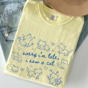 May include: Butter-colored t-shirt with a crew neck, featuring blue cartoon cat illustrations and the text "sorry i'm late, i saw a cat." The shirt is folded, with a hat and jeans in the background.