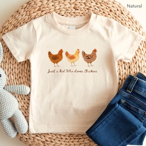 May include: Cream-colored t-shirt featuring three watercolor chickens in shades of brown and yellow. The text "Just a Kid Who Loves Chickens" is printed below the chickens. Dark blue denim jeans are folded next to the shirt.