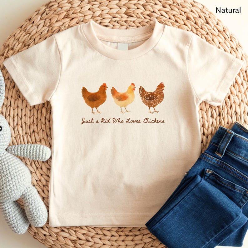 May include: Cream-colored t-shirt featuring three watercolor chickens in shades of brown and yellow. The text "Just a Kid Who Loves Chickens" is printed below the chickens. Dark blue denim jeans are folded next to the shirt.