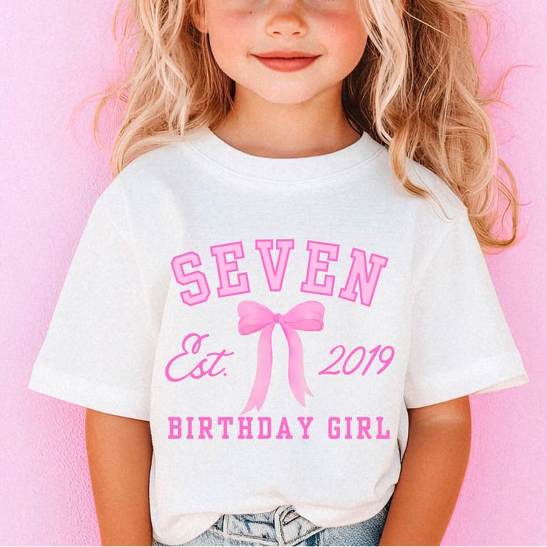 Peut inclure: T-shirt blanc avec l'inscription rose "SEVEN Est. 2019 BIRTHDAY GIRL". Le motif comprend un n&oelig;ud rose. Le t-shirt est &agrave; manches courtes.