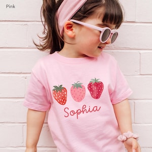 May include: Pink t-shirt featuring three strawberry illustrations and the name "Sophia" in a cursive font. The strawberries vary in color and detail. The word "Pink" is in the top left corner.