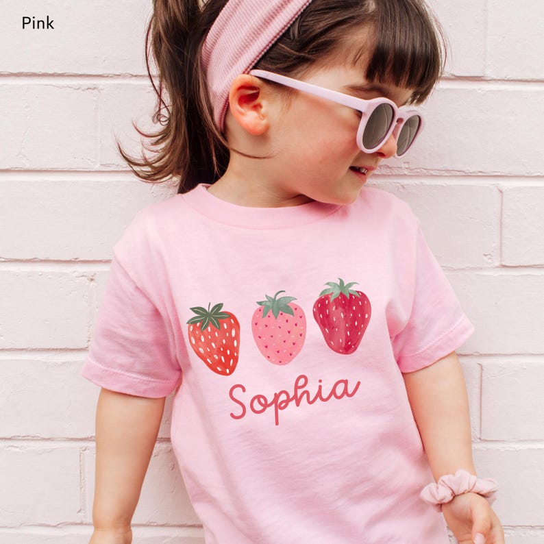 May include: Pink t-shirt featuring three strawberry illustrations and the name "Sophia" in a cursive font. The strawberries vary in color and detail. The word "Pink" is in the top left corner.
