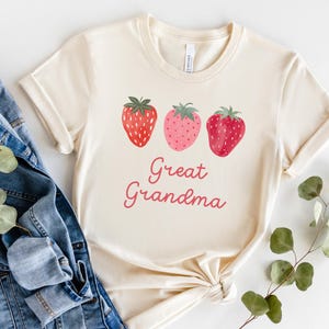 May include: Cream-colored t-shirt featuring three watercolor strawberries in shades of red and pink. The words "Great Grandma" are written in a cursive font below the strawberries. The shirt is folded and styled with a denim jacket.