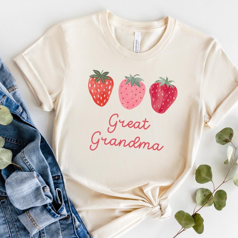 May include: Cream-colored t-shirt featuring three watercolor strawberries in shades of red and pink. The words "Great Grandma" are written in a cursive font below the strawberries. The shirt is folded and styled with a denim jacket.