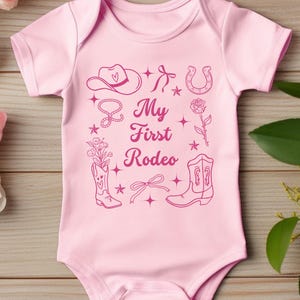 May include: Pink baby onesie with a pink graphic design of a cowboy hat, horseshoe, boots, lasso, rose, and stars. The text "My First Rodeo" is printed on the onesie.