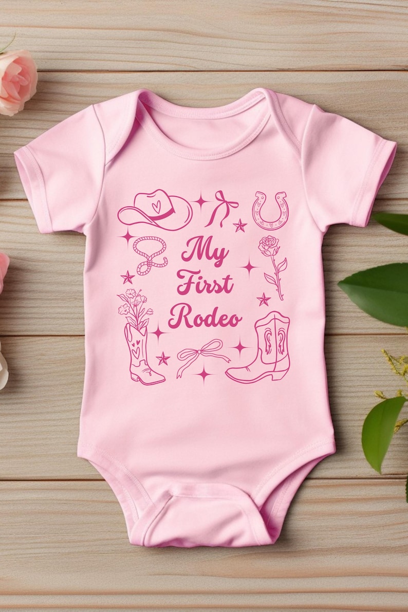 May include: Pink baby onesie with a pink graphic design of a cowboy hat, horseshoe, boots, lasso, rose, and stars. The text "My First Rodeo" is printed on the onesie.