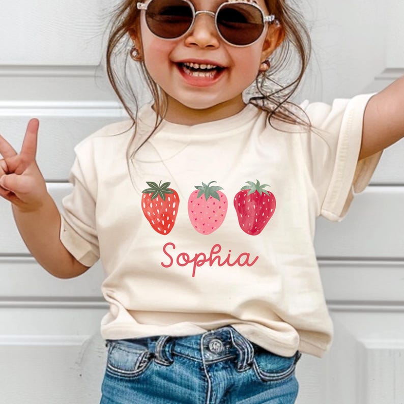 May include: A cream-colored t-shirt featuring three strawberry illustrations in shades of red and pink. The name "Sophia" is printed in a cursive font below the strawberries. The child is wearing blue jeans and sunglasses.