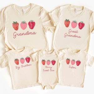 May include: A collection of cream-colored t-shirts and a onesie, each adorned with a strawberry design and a family-related title. The shirts feature the words "Grandma", "Great Grandma", "Big Brother", and "Sister", while the onesie says "Berry Sweet One".