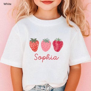 May include: White t-shirt featuring three strawberry illustrations in shades of red and pink. The name "Sophia" is written in a cursive font below the strawberries. The shirt is a short-sleeved design.