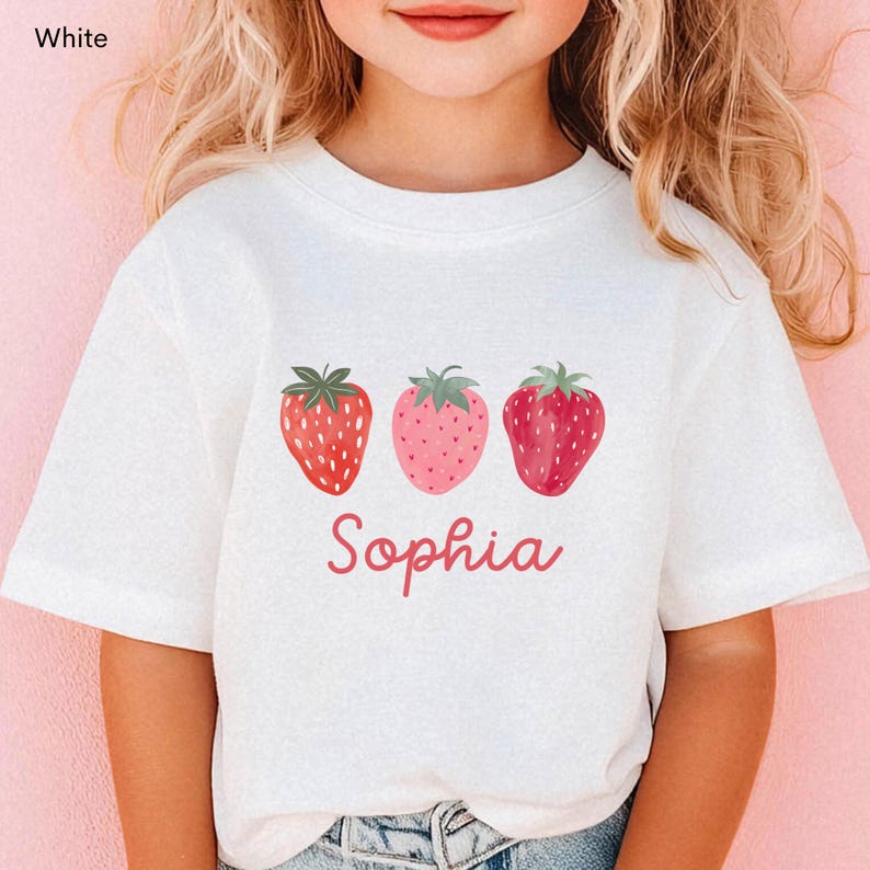 May include: White t-shirt featuring three strawberry illustrations in shades of red and pink. The name "Sophia" is written in a cursive font below the strawberries. The shirt is a short-sleeved design.