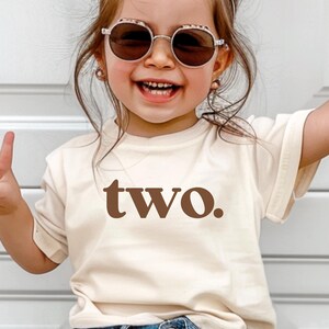 May include: A toddler wearing a light brown t-shirt with the word "two" printed in brown. The toddler is wearing blue jeans and round sunglasses.