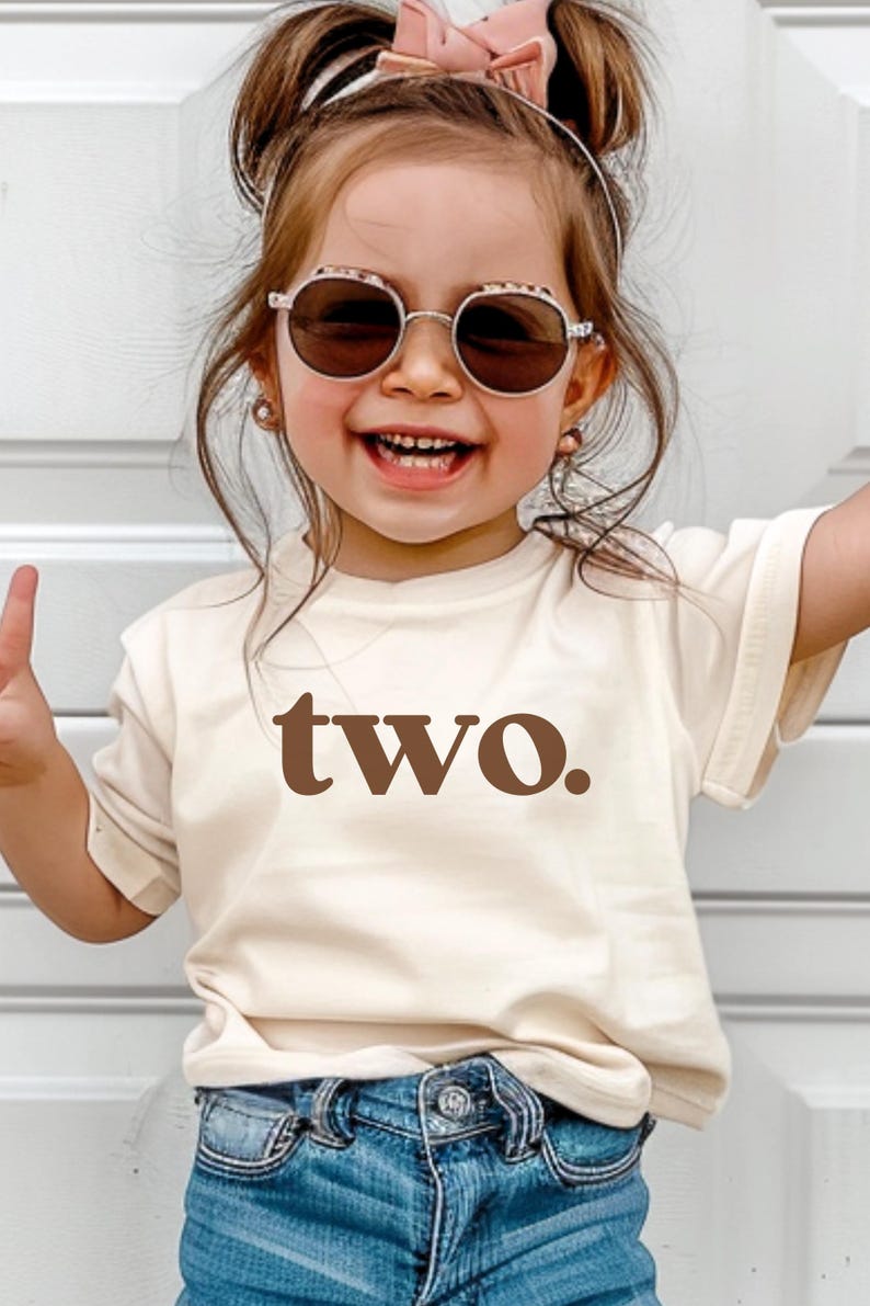 May include: A toddler wearing a light brown t-shirt with the word "two" printed in brown. The toddler is wearing blue jeans and round sunglasses.