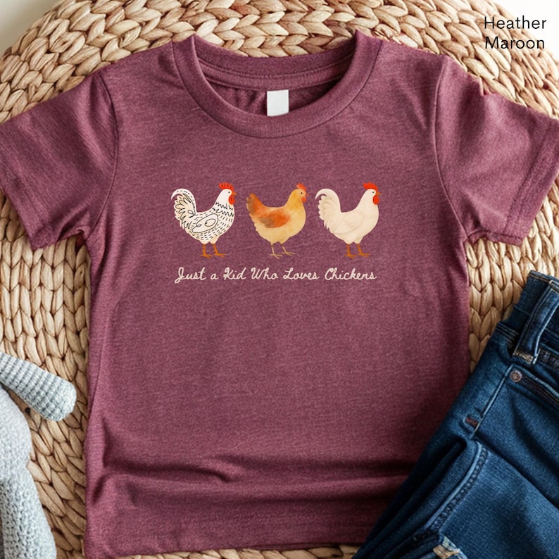 May include: Heather maroon t-shirt featuring three cartoon chickens in a row. The chickens are white, orange, and white with red accents. The text "Just a Kid Who Loves Chickens" is below the chickens.