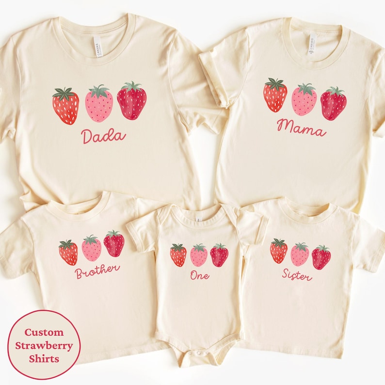 May include: Cream-colored family t-shirts and a onesie with a strawberry design. The shirts have the words "Dada", "Mama", "Brother", "One", and "Sister" printed below the strawberries. A circular graphic reads "Custom Strawberry Shirts".