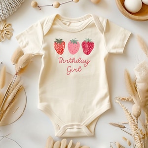 May include: Cream-colored baby bodysuit with three strawberry illustrations and the text "Birthday Girl" in pink. The strawberries vary in shades of red and pink, with green tops. The bodysuit is made of soft material and is designed for a baby.