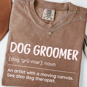 May include: Espresso-colored t-shirt with the words "DOG GROOMER" in large, light pink letters. The shirt includes a definition and the phrase "An artist with a moving canvas." A silver necklace with a small pendant is on the shirt.