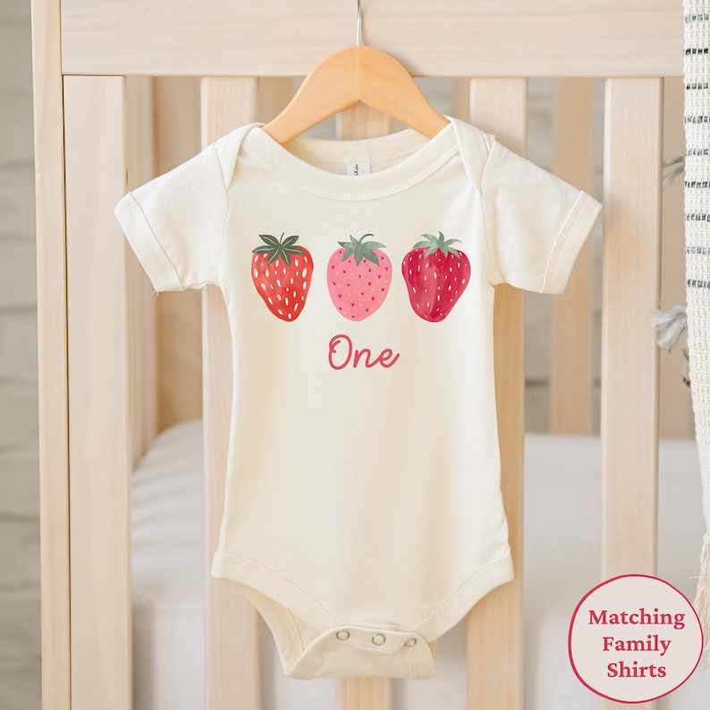 May include: Cream-colored baby bodysuit with three strawberry illustrations and the word "One" in red script. The strawberries are red and pink with green tops. The bodysuit is hanging on a wooden hanger.