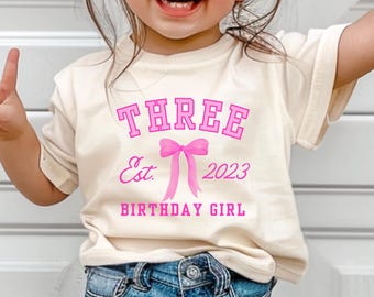 May include: Cream-colored t-shirt with the text "THREE Est. 2023 BIRTHDAY GIRL" in pink. The design includes a pink bow. The shirt is worn by a young child wearing sunglasses and blue jeans.