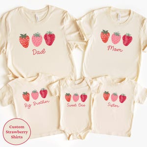 May include: A collection of cream-colored t-shirts and a onesie, each adorned with a strawberry design. The shirts are labeled "Dad," "Mom," "Big Brother," and "Sister," while the onesie says "Sweet One." A circular graphic reads "Custom Strawberry Shirts."