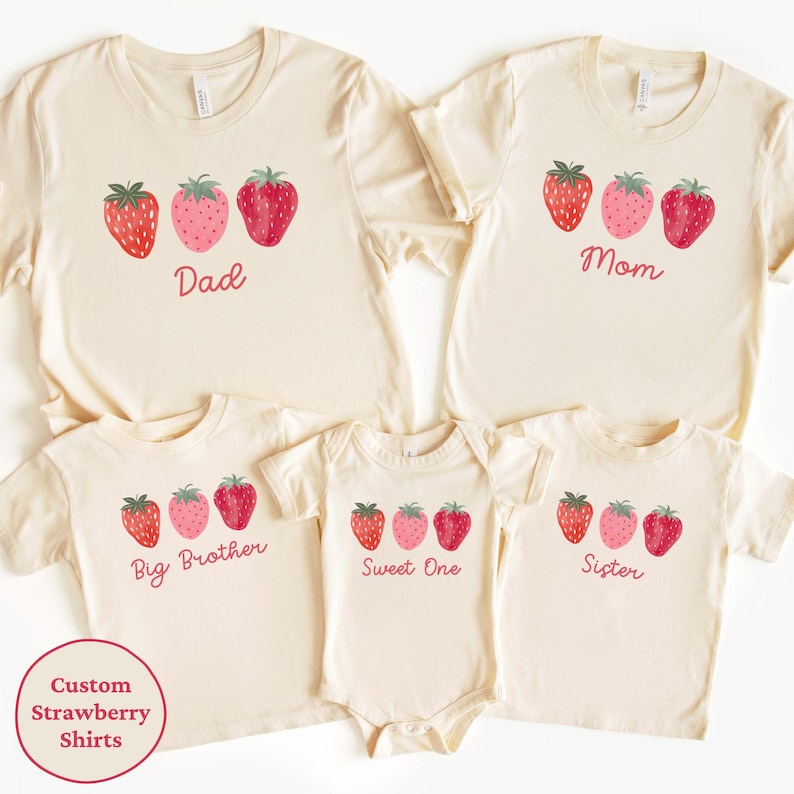 May include: A collection of cream-colored t-shirts and a onesie, each adorned with a strawberry design. The shirts are labeled "Dad," "Mom," "Big Brother," and "Sister," while the onesie says "Sweet One." A circular graphic reads "Custom Strawberry Shirts."