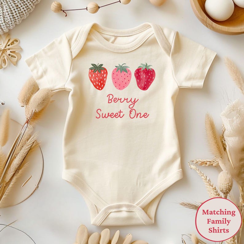 May include: Cream-colored baby onesie with three strawberry illustrations and the text "Berry Sweet One" in pink. The strawberries are red and pink with green tops. The onesie is short-sleeved and has a round neckline.