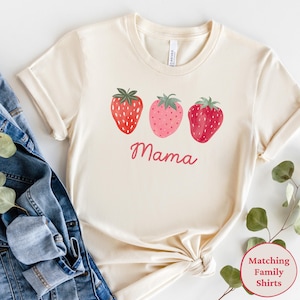 May include: Cream-colored t-shirt featuring three watercolor strawberries in shades of red and pink. The word "Mama" is written in cursive below the strawberries. The shirt is styled with a denim jacket and greenery.
