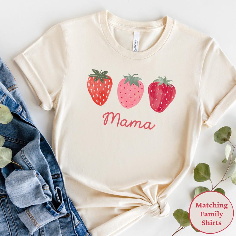 May include: Cream-colored t-shirt featuring three watercolor strawberries in shades of red and pink. The word "Mama" is written in cursive below the strawberries. The shirt is styled with a denim jacket and greenery.
