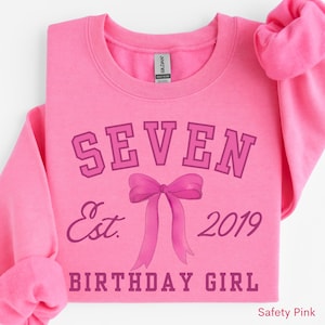 Peut inclure: Un sweat-shirt rose avec le mot &laquo;&nbsp;SEVEN&nbsp;&raquo; au-dessus d'un motif de n&oelig;ud et les mots &laquo;&nbsp;Est. 2019 BIRTHDAY GIRL&nbsp;&raquo; dans une police rose plus fonc&eacute;e. Le sweat-shirt est de couleur rose uni.