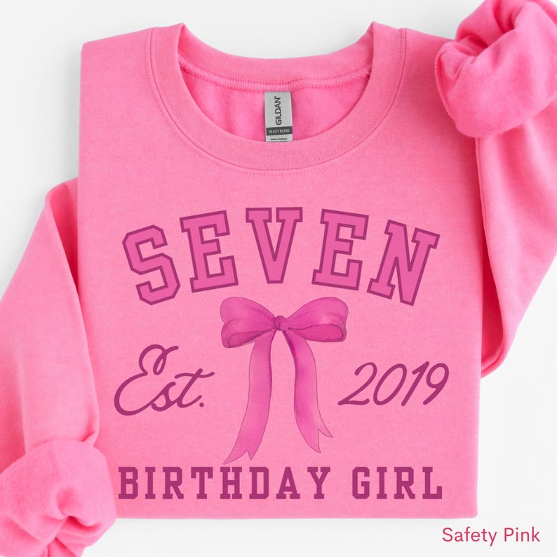 Peut inclure: Un sweat-shirt rose avec le mot &laquo;&nbsp;SEVEN&nbsp;&raquo; au-dessus d'un motif de n&oelig;ud et les mots &laquo;&nbsp;Est. 2019 BIRTHDAY GIRL&nbsp;&raquo; dans une police rose plus fonc&eacute;e. Le sweat-shirt est de couleur rose uni.