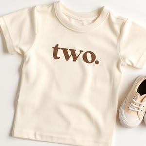 May include: A cream colored short-sleeved t-shirt with the word "two" printed in brown on the front.