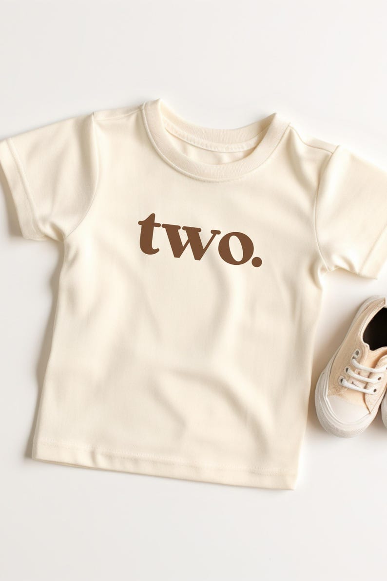 May include: A cream colored short-sleeved t-shirt with the word "two" printed in brown on the front.