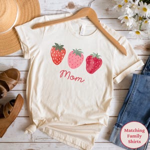May include: Cream-colored t-shirt featuring three strawberry illustrations in red and pink, with green leaves. The word "Mom" is written in red script below the strawberries. The shirt is displayed on a wooden hanger.