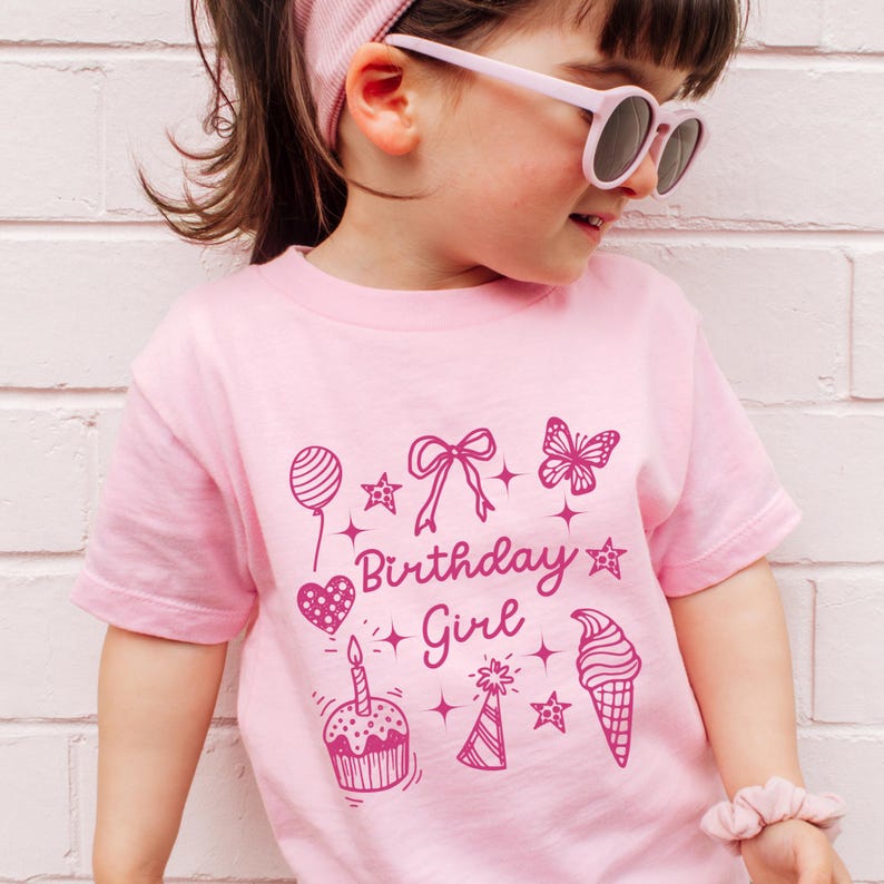 May include: Pink t-shirt with a pink graphic design that says "Birthday Girl" and includes a cupcake, ice cream cone, balloon, butterfly, and stars.