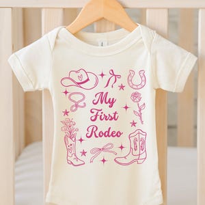 May include: A white baby onesie with a pink graphic design of a cowboy hat, horseshoe, boots, and a lasso. The text "My First Rodeo" is printed in pink on the onesie.