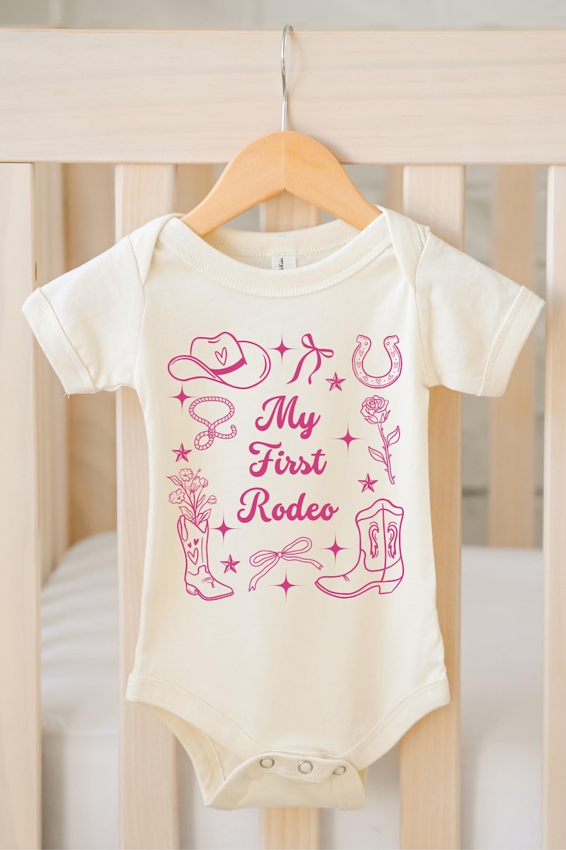 May include: A white baby onesie with a pink graphic design of a cowboy hat, horseshoe, boots, and a lasso. The text "My First Rodeo" is printed in pink on the onesie.