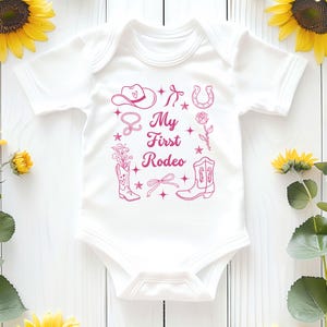 May include: White baby onesie with pink outline illustrations of a cowboy hat, boots, horseshoe, and flowers. The text "My First Rodeo" is printed in pink.
