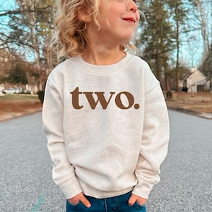 May include: A beige sweatshirt with the word "two." in brown lettering. The sweatshirt is worn by a young child with blonde hair and blue jeans. The child is standing on a paved road.