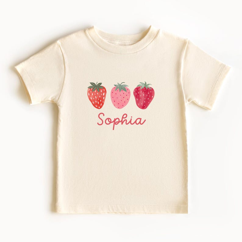 May include: Cream-colored t-shirt featuring three watercolor strawberries in shades of red and pink. The name "Sophia" is written in a matching pink script below the strawberries. The shirt has short sleeves and a crew neck.