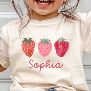 May include: Cream-colored t-shirt featuring three watercolor strawberries in shades of red and pink. The name "Sophia" is written in a cursive font below the strawberries. The shirt is worn by a child.