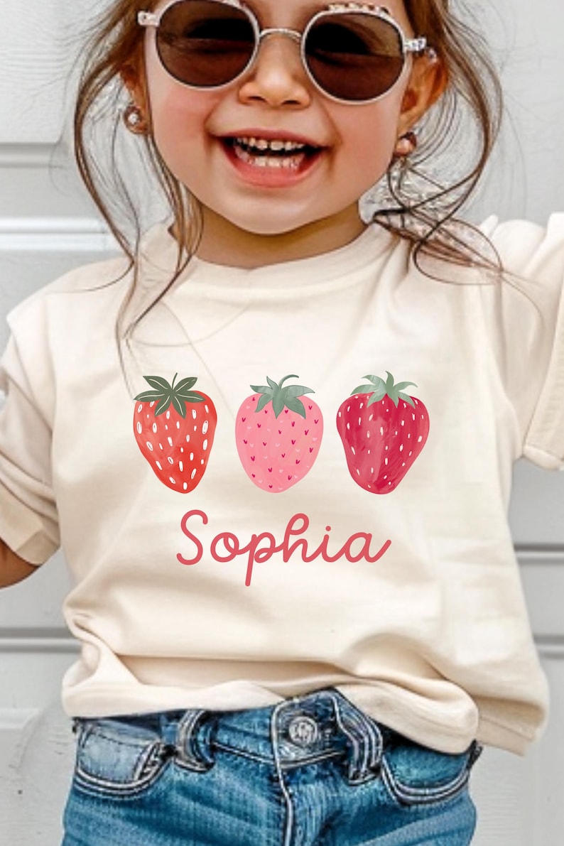 May include: Cream-colored t-shirt featuring three watercolor strawberries in shades of red and pink. The name "Sophia" is written in a cursive font below the strawberries. The shirt is worn by a child.