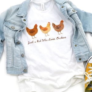 May include: White t-shirt with three watercolor chickens and the text "Just a Kid Who Loves Chickens." A light blue denim jacket is draped over the shirt. Yellow sneakers are visible in the bottom right corner.